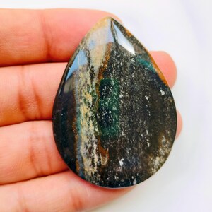 May include: A teardrop-shaped gemstone with a polished, glossy surface. The stone features a mix of colours, including dark green, brown, and blue, with white speckles. The gemstone is held in a hand.