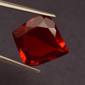 Peut inclure: Une pierre précieuse rouge foncé à facettes, tenue par une pince argentée. Le bijou a une forme carrée avec un bord légèrement incurvé et de multiples surfaces réfléchissantes. Le fond est gris foncé.