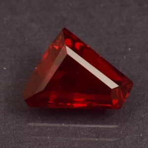 May include: A faceted, triangular gemstone in a deep red colour. The gem has multiple facets that reflect light, creating a brilliant shine. The gemstone is set against a dark background.