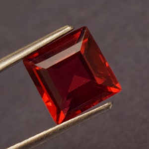 Op de afbeelding: Een gefacetteerde, vierkant geslepen, dieprode edelsteen, vastgehouden door een zilveren pincet. De edelsteen heeft een briljant, reflecterend oppervlak en een rijke, verzadigde kleur. De edelsteen heeft een vierkante vorm met schuine randen.