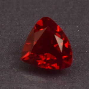 May include: A faceted, triangular-shaped, deep red gemstone. The gem has a polished surface that reflects light, creating bright highlights. The stone is set against a dark gray background.