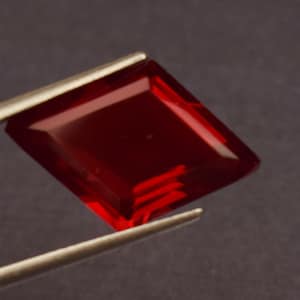 May include: A faceted, rectangular-cut, deep red gemstone is held by silver tweezers. The gem has a high degree of clarity and a polished surface, reflecting light. The gemstone's shape is a modified emerald cut, with beveled edges.