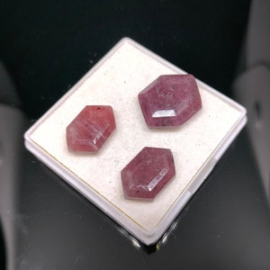 May include: Three faceted, reddish-pink gemstones with a hexagonal shape are displayed on a white surface. The gems have a polished appearance, reflecting light. The background is dark, highlighting the gems.