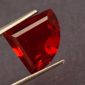 May include: A faceted, semi-circular, deep red gemstone held by silver tweezers. The gem has a polished, reflective surface with sharp, geometric edges. The gem's color is a rich, saturated red, with a slight translucence.