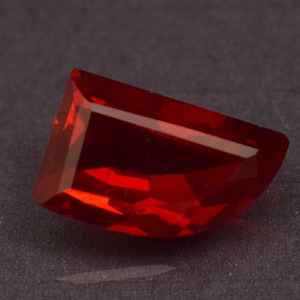 Peut inclure: Une pierre précieuse taillée en forme rectangulaire, d'un rouge profond et vibrant. Les surfaces polies de la gemme reflètent la lumière, mettant en valeur sa clarté et sa brillance. La pierre est posée sur un fond sombre.