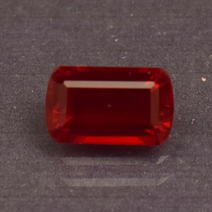 May include: A rectangular, faceted, deep red gemstone with rounded corners. The gem is highly polished, reflecting light and showcasing its rich color. The stone is set against a dark gray background, highlighting its brilliance.