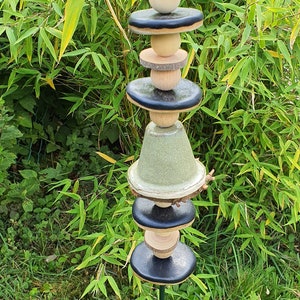 Könnte beinhalten: Ein Keramik-Gartenpfahl mit einem weißen, grünen und schwarzen Design. Der Pfahl besteht aus mehreren übereinander gestapelten Teilen, darunter eine weiße Keramikglocke, eine grüne Keramikglocke und schwarze und hölzerne Scheiben. Der Pfahl ist in einem grasbewachsenen Bereich in den Boden gesteckt.