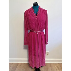 May include: A long, fuchsia-colored dress with a pleated design. The dress features long sleeves, a V-neckline with ruffled trim, and a matching belt. The dress is displayed on a mannequin.