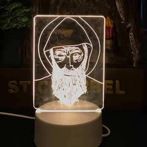 Könnte beinhalten: Weißes Acryl-Nachtlicht mit einer weißen Umrandung eines Mannes mit Bart und Kapuze. Das Nachtlicht steht auf einem weißen Sockel und hat ein Kabel.