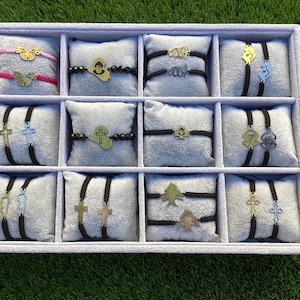 May include: A white display case with 10 compartments, each containing two black string bracelets with different gold charms. The charms include a cross, a butterfly, a tree, a heart, and a geometric design.