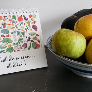 May include: A white calendar with a colorful illustration of fruits and vegetables. The calendar says "C'est de saison... et d'ici!" and is sitting on a dark surface next to a bowl of fruit. The bowl contains a green apple, a yellow apple, a yellow pear, and a purple eggplant.