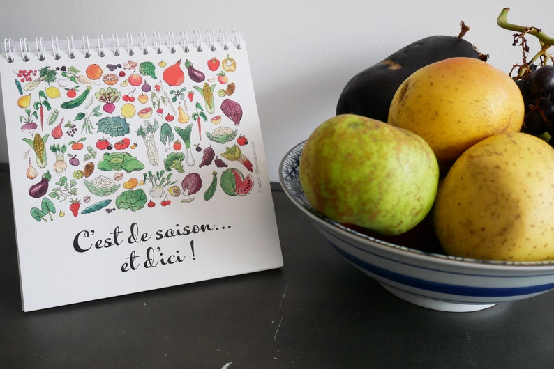 May include: A white calendar with a colorful illustration of fruits and vegetables. The calendar says "C'est de saison... et d'ici!" and is sitting on a dark surface next to a bowl of fruit. The bowl contains a green apple, a yellow apple, a yellow pear, and a purple eggplant.
