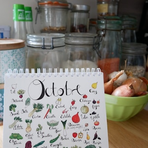 May include: A white calendar with the word "October" written in black cursive. The calendar features a colorful illustration of various fruits and vegetables, including beets, broccoli, carrots, cauliflower, celery, onions, and tomatoes. The calendar is sitting on a wooden surface.