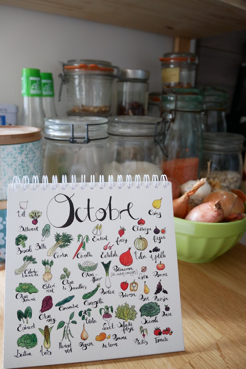 May include: A white calendar with the word "October" written in black cursive. The calendar features a colorful illustration of various fruits and vegetables, including beets, broccoli, carrots, cauliflower, celery, onions, and tomatoes. The calendar is sitting on a wooden surface.