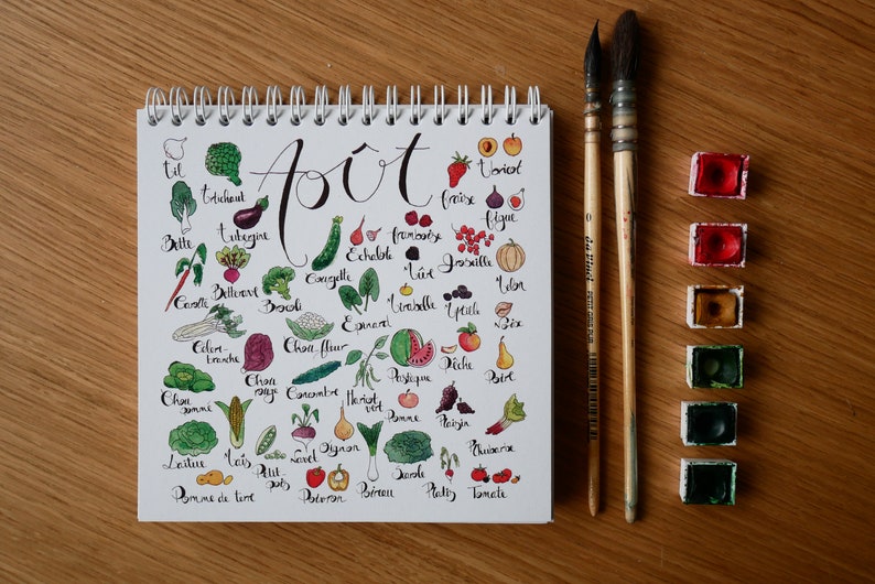 May include: A white notebook with a spiral binding and a handwritten list of fruits and vegetables in French. The notebook is open to a page titled "Aout" and features illustrations of each item. The notebook is on a wooden surface with a set of watercolor paints.