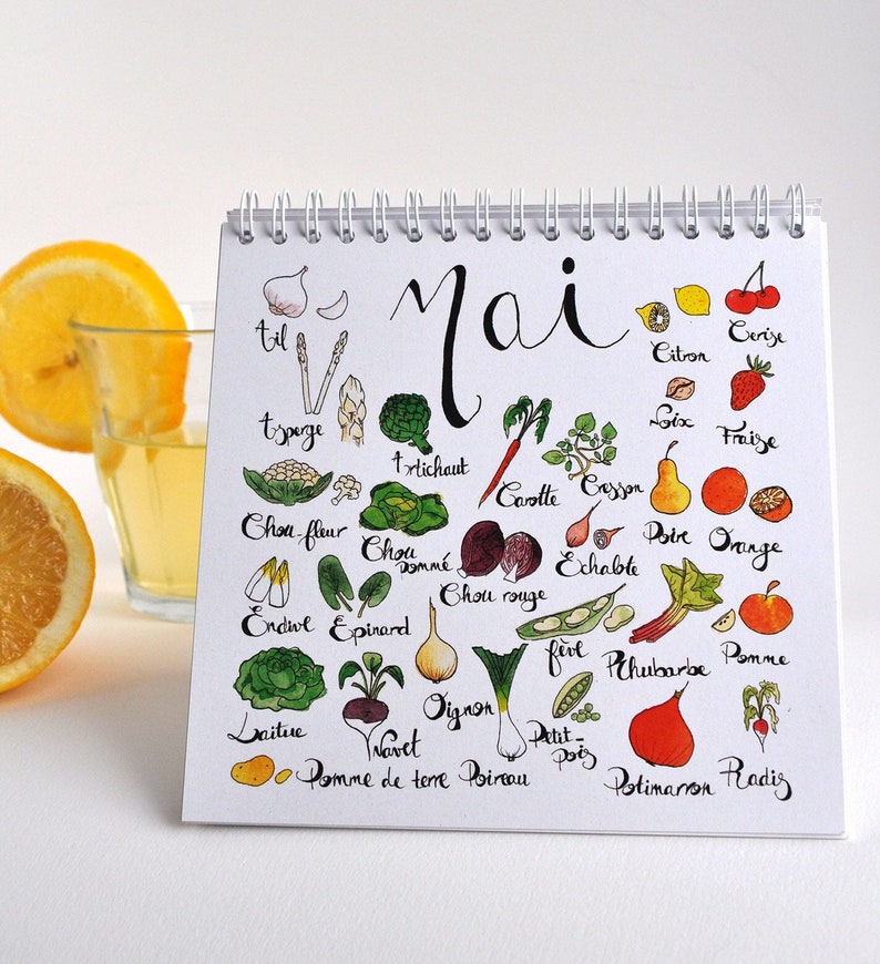 May include: A white calendar with the word "Mai" (May in German) written in black cursive. The calendar lists various fruits and vegetables that are in season during the month of May. The fruits and vegetables are illustrated with hand-drawn images.