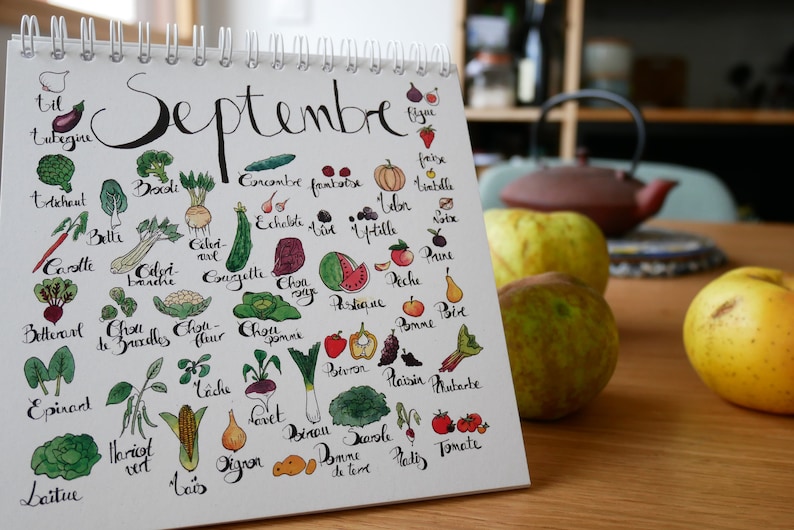 May include: A white calendar with the word "Septembre" written in black cursive. The calendar lists various fruits and vegetables in French, with hand-drawn illustrations of each item.