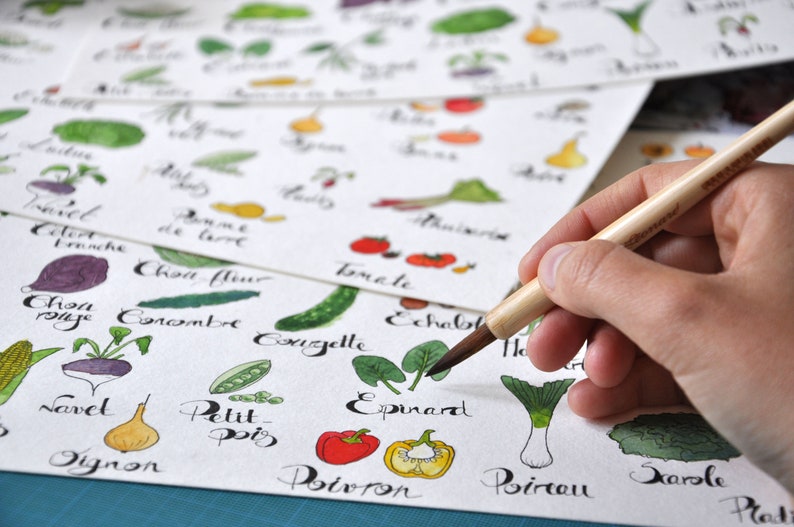 May include: A hand holding a paintbrush over a sheet of paper with hand-drawn illustrations of vegetables and their names in French. The vegetables include a carrot, onion, tomato, eggplant, and spinach.