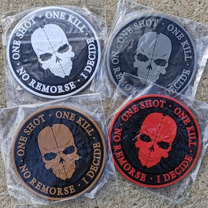 Puede incluir: Cuatro parches redondos negros con un diseño de calavera y huesos cruzados blancos. Los parches tienen el texto "One Shot. One Kill. No Remorse. I Decide." alrededor de la calavera. Los parches son de diferentes colores: blanco, gris, marrón y rojo.