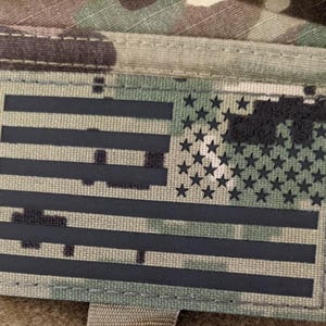 Può includere: Toppa rettangolare con un design stilizzato della bandiera americana in bianco e nero. La bandiera presenta strisce e stelle nere su uno sfondo mimetico. La toppa è probabilmente realizzata in tessuto resistente.