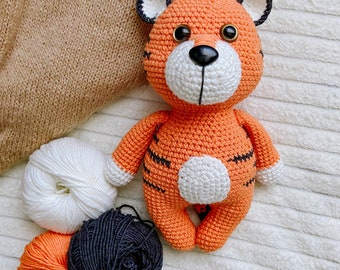 May include: A handmade crocheted tiger doll in orange and black, with white details and black stripes. The doll has large eyes and is next to three balls of yarn in white, orange, and black. The tiger has a white belly and a small white tail.