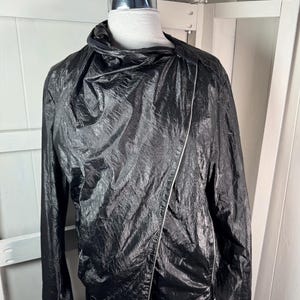 Pode incluir: Casaco preto com acabamento brilhante e texturizado. O casaco apresenta um zíper assimétrico e uma gola alta drapeada. Uma faixa branca acentua o zíper. O casaco é exibido em um manequim.