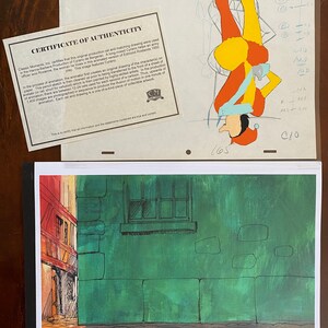 May include: An original animation cel from the Hanna-Barbera production of Cyrano de Bergerac, featuring a colorful jester character. The image includes a certificate of authenticity and a background scene of a building with a green wall.