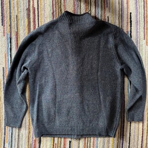 May include: A gray knit sweater with a high collar and ribbed cuffs and hem. The sweater has a textured pattern and a zipper closure. The background is a striped rug.