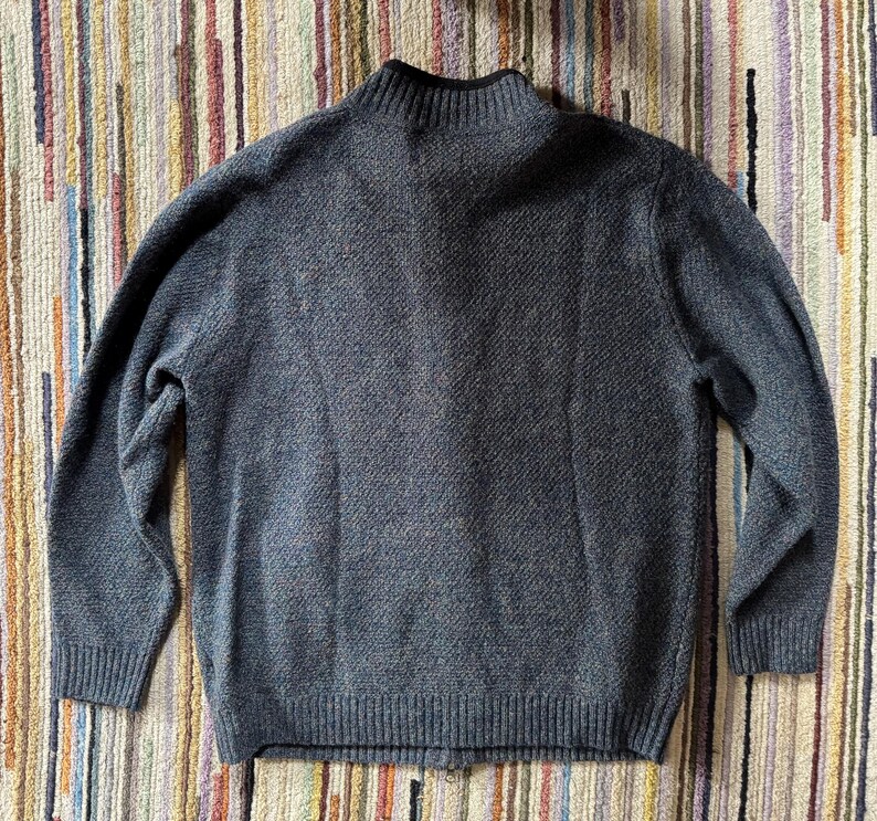 May include: A gray knit sweater with a high collar and ribbed cuffs and hem. The sweater has a textured pattern and a zipper closure. The background is a striped rug.