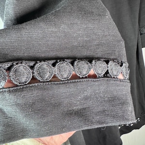 May include: Close-up of a dark gray garment sleeve with a decorative, scalloped lace trim. The fabric appears to be a soft, textured material. The sleeve is partially visible, showcasing the intricate detailing of the lace.