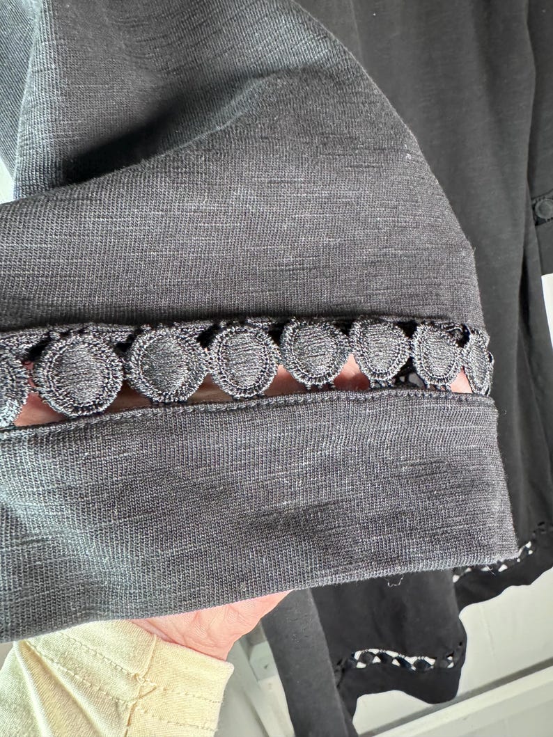 May include: Close-up of a dark gray garment sleeve with a decorative, scalloped lace trim. The fabric appears to be a soft, textured material. The sleeve is partially visible, showcasing the intricate detailing of the lace.