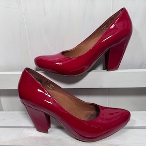 Puede incluir: Un par de zapatos de tacón alto rojos brillantes. Los zapatos tienen una punta redondeada y un tacón grueso. Los tacones miden aproximadamente 7.6 cm de alto. Los zapatos se exhiben sobre una superficie blanca.