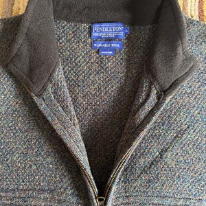 May include: A close-up of a blue, green, and brown wool zip-up jacket with a black collar. The jacket has a zipper closure and a label that reads "PENDLETON WASHABLE WOOL".