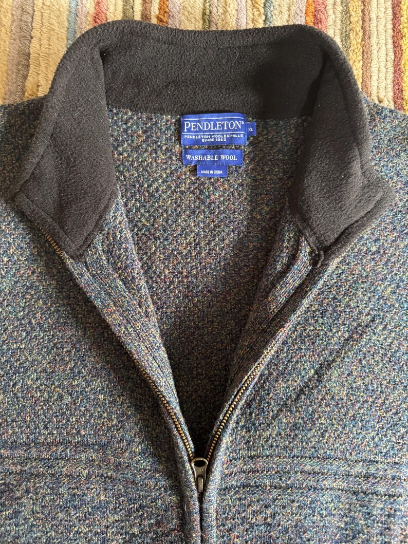 May include: A close-up of a blue, green, and brown wool zip-up jacket with a black collar. The jacket has a zipper closure and a label that reads "PENDLETON WASHABLE WOOL".