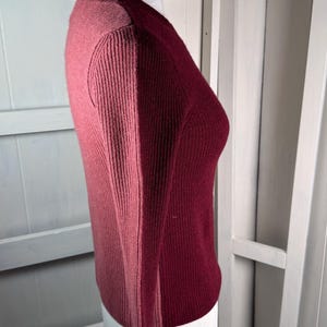 Puede incluir: Un suéter de manga larga de dos tonos en tonos rosa y burdeos. El suéter de punto acanalado se exhibe en un maniquí, mostrando su diseño ajustado. El estilo de bloques de color agrega un toque moderno a la prenda de punto clásica.