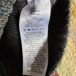 May include: Close-up of a white clothing tag with black text. The tag provides fabric composition details: 100% Shetland wool and 100% polyester. Washing instructions are also included. The tag is attached to a dark-colored knitted garment.