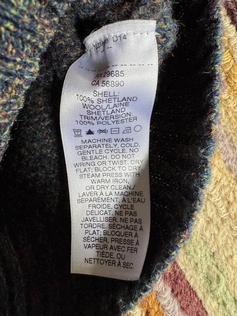 May include: Close-up of a white clothing tag with black text. The tag provides fabric composition details: 100% Shetland wool and 100% polyester. Washing instructions are also included. The tag is attached to a dark-colored knitted garment.