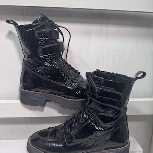 May include: Pair of black patent leather combat boots with laces and buckle straps. The boots have a chunky sole with a brown trim and a pull tab at the back. The boots are displayed on a white wooden surface.