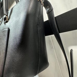 May include: A black leather tote bag with a gold chain detail along the top edge. The bag has two handles and a long, detachable strap. The leather appears smooth and the bag has a rectangular shape.