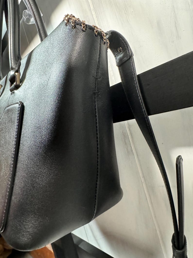 May include: A black leather tote bag with a gold chain detail along the top edge. The bag has two handles and a long, detachable strap. The leather appears smooth and the bag has a rectangular shape.
