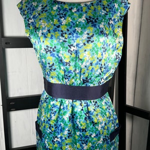 May include: A sleeveless dress with a floral pattern in shades of blue, green, and white. The dress has a navy blue waist band and pockets. The dress is displayed on a mannequin.