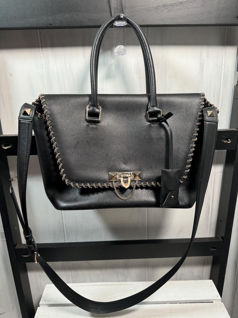 May include: Black leather handbag with gold-tone chain and stud accents. Features a top handle, detachable shoulder strap, and a small attached pouch. The bag has a rectangular shape with a flap closure and a gold-tone lock.