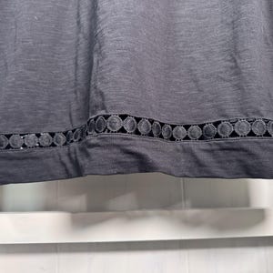 May include: A dark gray top with a decorative, circular lace trim along the bottom edge. The fabric appears to be a soft knit material. The trim is a darker shade of gray, creating a subtle contrast.