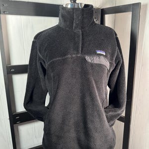 May include: Black fleece pullover with a high collar and a small chest pocket. The pullover has a button closure at the neck and a small logo patch on the chest pocket. The material appears soft and textured.