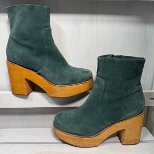 May include: A pair of teal suede ankle boots with wooden platform soles and block heels. The boots have a side zipper and a rounded toe. The wooden platform and heel are a light brown color.