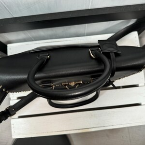 May include: Black leather handbag with a top handle and a detachable shoulder strap. The bag features gold-toned hardware, including a clasp closure and chain detailing along the edges. The bag is sitting on a white surface.