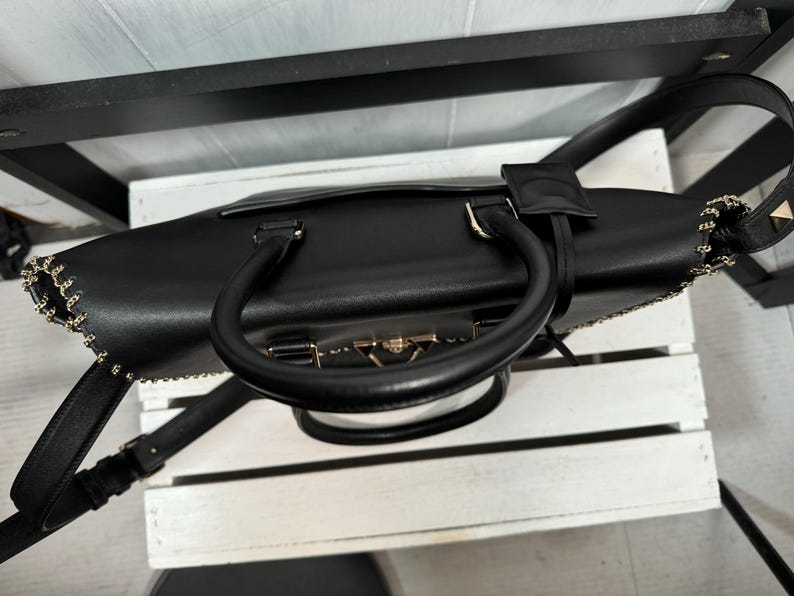 May include: Black leather handbag with a top handle and a detachable shoulder strap. The bag features gold-toned hardware, including a clasp closure and chain detailing along the edges. The bag is sitting on a white surface.