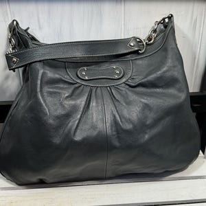 Pode incluir: Bolsa de ombro de couro preta com alças de corrente prateadas e uma alça de couro larga. A bolsa tem um design franzido e um pequeno patch decorativo de couro com costura branca. A bolsa está em uma superfície de madeira branca.