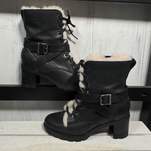 May include: A pair of black leather boots with a lace-up design and a buckle strap. The boots have a faux fur lining and a block heel. The boots are displayed on a white wooden surface.