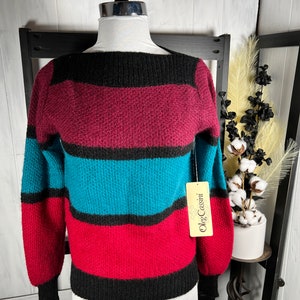 Puede incluir: Un suéter a rayas en negro, burdeos, turquesa y rojo. El suéter tiene cuello barco y mangas largas. La etiqueta dice "Oleg Cassini". El suéter se exhibe en un maniquí.